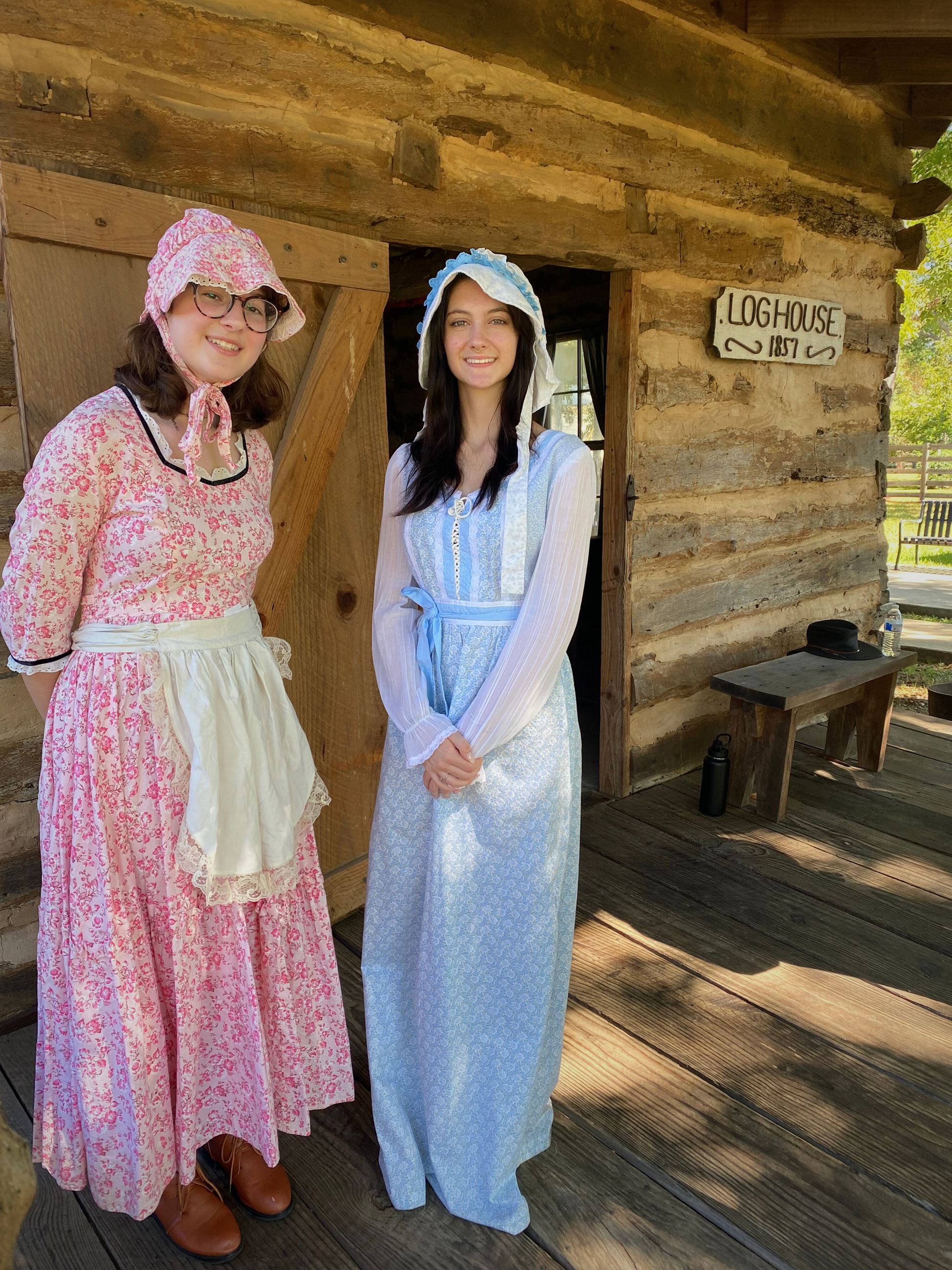picture of women in period attire in from of a loghouse